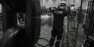 Mitglied der MER Klang Crew beim Aufbau von Veranstaltungsequipment, erkennbar am'MERCREW'-Schriftzug auf dem T-Shirt des Technikers, der professionelle Event-Vorbereitungen in einer Messe-Halle durchführt.